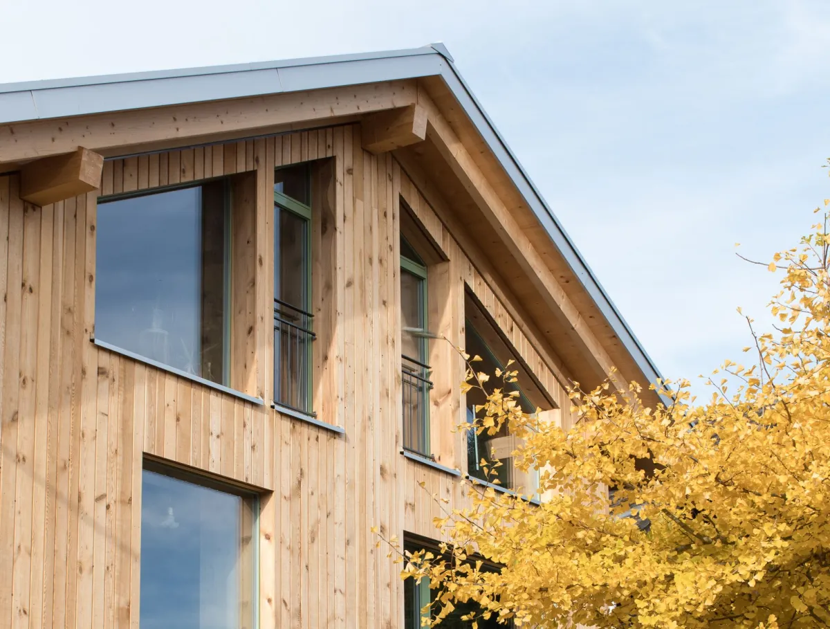 Aussenansicht eines mehrgeschossigen Wohnhauses in moderner Holzbauweise mit senkrechter Holzfassade und grossen Fenstern. Vor dem Gebäude steht ein Baum mit gelben Herbstblättern, der Teile der Fassade überragt. Der Eingangsbereich und der Garten mit Pflanzen und Pflasterflächen sind im Vordergrund sichtbar, bei hellem Tageslicht.