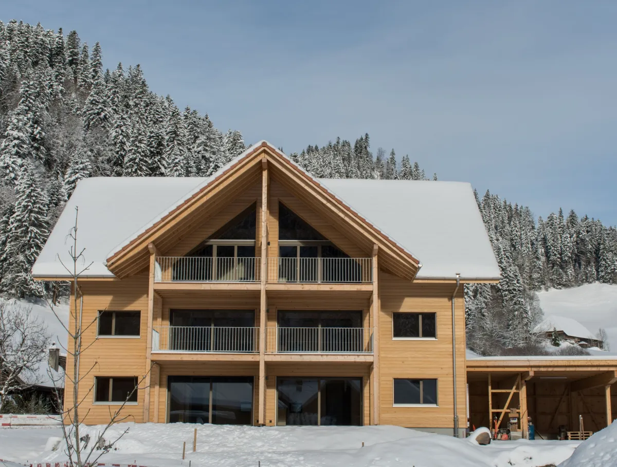 Aussenansicht eines neu gebauten Mehrfamilienhauses in Holzbauweise mit Satteldach und Balkonen im Winter. Das Gebäude steht in verschneiter Landschaft neben einem landwirtschaftlichen Anbau, umgeben von Hügeln und verschneiten Bäumen unter blauem Himmel.