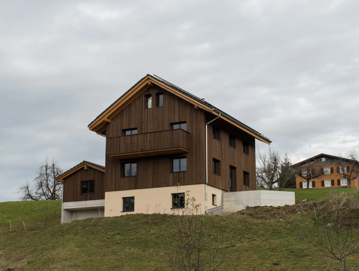 Neubau eines modernen Holzhauses am Hang mit dunkler Holzfassade, Balkon und grosser Fensterfront. Das Gebäude steht in ländlicher Umgebung mit Wiese und weiteren Häusern im Hintergrund unter bewölktem Himmel.