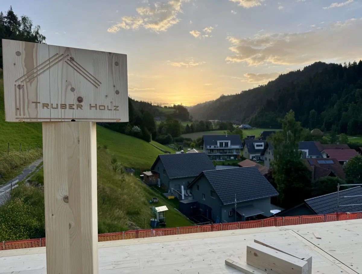 Zimmerleute montieren während der Aufrichtearbeiten eine vorgefertigte Holzkonstruktion auf einem Gebäude. Ein Kran hebt ein grosses Dachelement an seinen Platz, während mehrere Fachkräfte auf dem Dach die Montage begleiten.