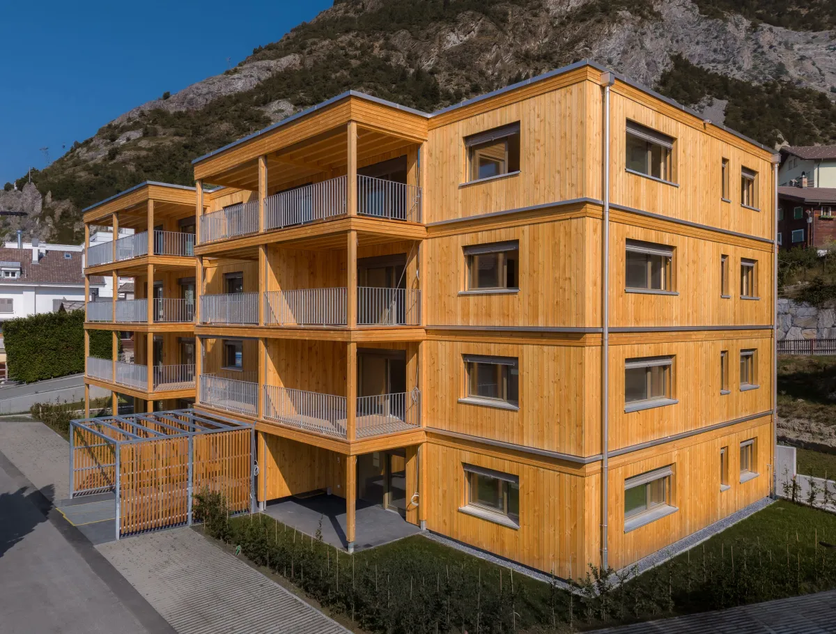 Aussenansicht eines modernen, viergeschossigen Mehrfamilienhauses in Holzbauweise vor einer felsigen Bergkulisse. Das Gebäude verfügt über eine vertikale Holzfassade sowie grosszügige, überdachte Balkone mit sichtbarer Holzkonstruktion und Metallgeländern.