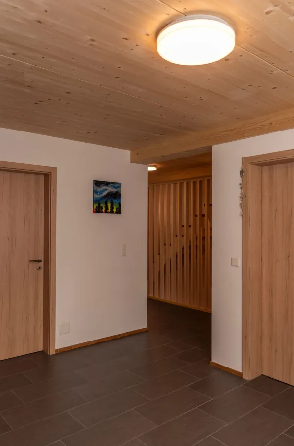 Aussenansicht eines neu errichteten Wohnhauses in Holzbauweise mit Satteldach und Balkonen, eingebettet in eine hügelige, ländliche Landschaft. Das Gebäude steht neben bestehenden landwirtschaftlichen Bauten. Im Vordergrund eine Wiese, im Hintergrund ein begrünter Hang mit Bäumen unter blauem Himmel.