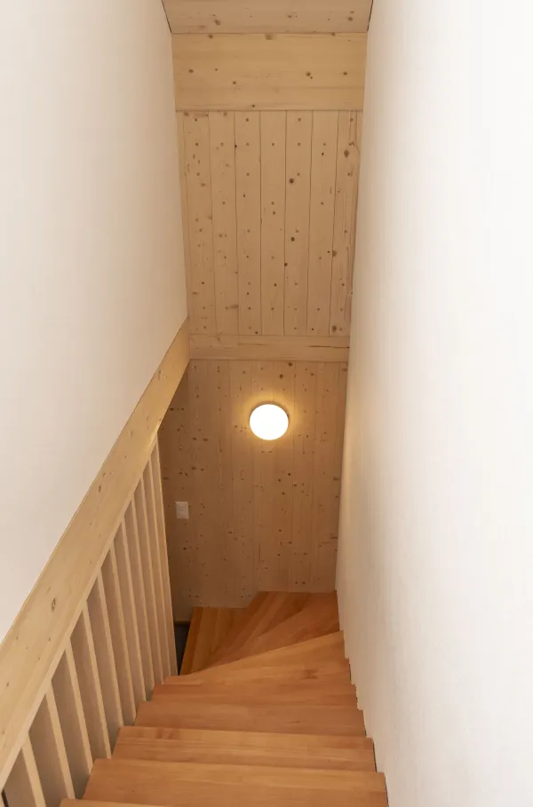 Aussenansicht eines neu errichteten Wohnhauses in Holzbauweise mit Satteldach und Balkonen, eingebettet in eine hügelige, ländliche Landschaft. Das Gebäude steht neben bestehenden landwirtschaftlichen Bauten. Im Vordergrund eine Wiese, im Hintergrund ein begrünter Hang mit Bäumen unter blauem Himmel.