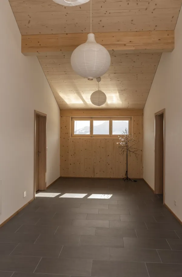 Aussenansicht eines neu errichteten Wohnhauses in Holzbauweise mit Satteldach und Balkonen, eingebettet in eine hügelige, ländliche Landschaft. Das Gebäude steht neben bestehenden landwirtschaftlichen Bauten. Im Vordergrund eine Wiese, im Hintergrund ein begrünter Hang mit Bäumen unter blauem Himmel.