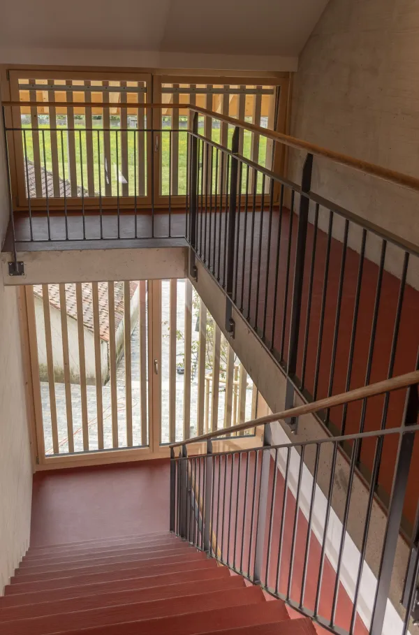 Innenansicht eines hellen Wohnraums in Holzbauweise mit sichtbarer Holzdecke und Holzboden. Grosse, bodentiefe Schiebetüren aus Glas führen auf einen überdachten Balkon mit Holzgeländer. Durch die Fenster fällt viel Tageslicht ein und ermöglicht den Blick nach draussen.