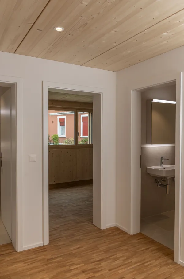 Innenansicht eines hellen Wohnraums in Holzbauweise mit sichtbarer Holzdecke und Holzboden. Grosse, bodentiefe Schiebetüren aus Glas führen auf einen überdachten Balkon mit Holzgeländer. Durch die Fenster fällt viel Tageslicht ein und ermöglicht den Blick nach draussen.