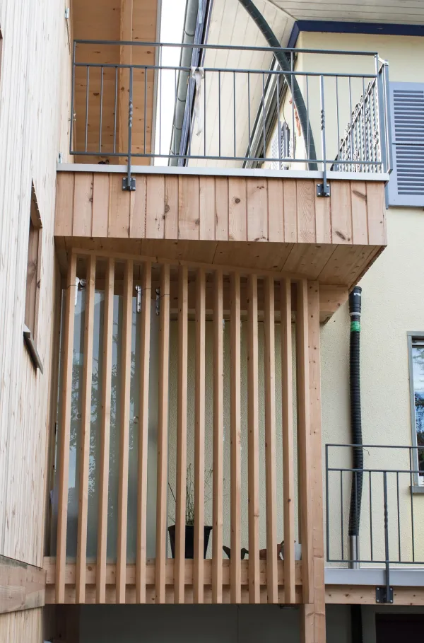 Aussenansicht eines mehrgeschossigen Wohnhauses in moderner Holzbauweise mit senkrechter Holzfassade und grossen Fenstern. Vor dem Gebäude steht ein Baum mit gelben Herbstblättern, der Teile der Fassade überragt. Der Eingangsbereich und der Garten mit Pflanzen und Pflasterflächen sind im Vordergrund sichtbar, bei hellem Tageslicht.