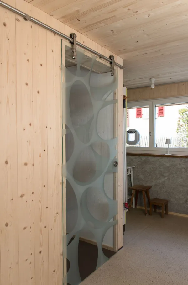 Innenansicht eines offenen Wohn- und Essbereichs in Holzbauweise. Decke und Wände sind mit hellem Holz verkleidet. In der Mitte steht eine Kücheninsel mit offenem Regal und Geschirr. Im Hintergrund befinden sich ein Esstisch mit Stühlen, ein Sitzbereich sowie grosse Fenster, die viel Tageslicht in den wohnlichen Raum lassen.