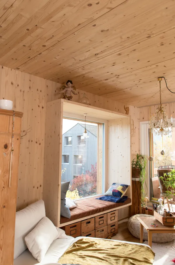 Innenansicht eines offenen Wohn- und Essbereichs in Holzbauweise. Decke und Wände sind mit hellem Holz verkleidet. In der Mitte steht eine Kücheninsel mit offenem Regal und Geschirr. Im Hintergrund befinden sich ein Esstisch mit Stühlen, ein Sitzbereich sowie grosse Fenster, die viel Tageslicht in den wohnlichen Raum lassen.