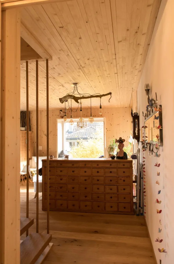 Innenansicht eines offenen Wohn- und Essbereichs in Holzbauweise. Decke und Wände sind mit hellem Holz verkleidet. In der Mitte steht eine Kücheninsel mit offenem Regal und Geschirr. Im Hintergrund befinden sich ein Esstisch mit Stühlen, ein Sitzbereich sowie grosse Fenster, die viel Tageslicht in den wohnlichen Raum lassen.