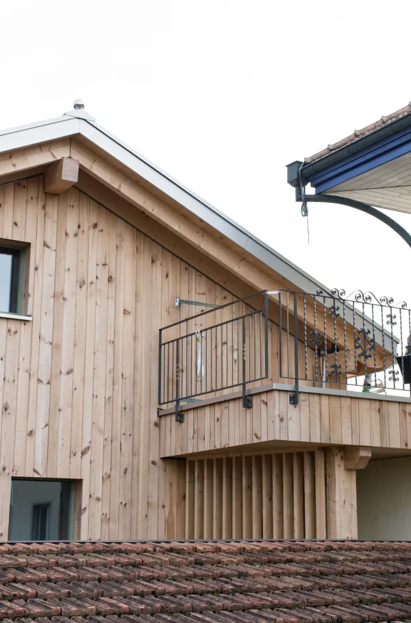 Aussenansicht eines mehrgeschossigen Wohnhauses in moderner Holzbauweise mit senkrechter Holzfassade und grossen Fenstern. Vor dem Gebäude steht ein Baum mit gelben Herbstblättern, der Teile der Fassade überragt. Der Eingangsbereich und der Garten mit Pflanzen und Pflasterflächen sind im Vordergrund sichtbar, bei hellem Tageslicht.