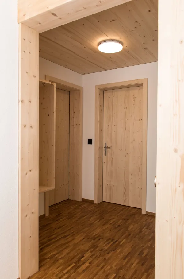 Heller Innenraum eines neu gebauten Holzhauses mit sichtbaren Holzbalken, offener Küche aus Holz, Treppenaufgang und grossen Fenstern mit Zugang zum Balkon. Der Raum wirkt modern, offen und natürlich durch die Kombination von Holz und klaren Linien.