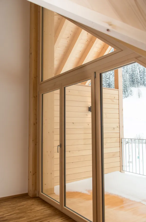 Heller Innenraum eines neu gebauten Holzhauses mit sichtbaren Holzbalken, offener Küche aus Holz, Treppenaufgang und grossen Fenstern mit Zugang zum Balkon. Der Raum wirkt modern, offen und natürlich durch die Kombination von Holz und klaren Linien.