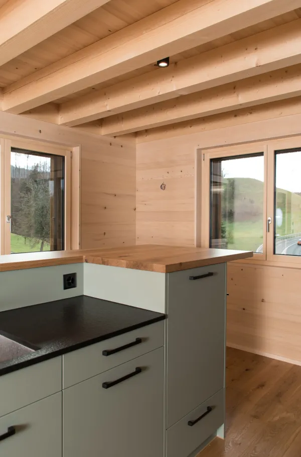 Innenansicht eines modernen Holzbaus mit sichtbaren Balken, natürlichen Holzoberflächen und massgefertigten Einbauten. Grosszügige Fensterflächen sorgen für viel Tageslicht und unterstreichen die warme, ruhige Wohnatmosphäre. Hochwertige Materialien und präzise Schreinerarbeiten prägen das Gesamtbild.