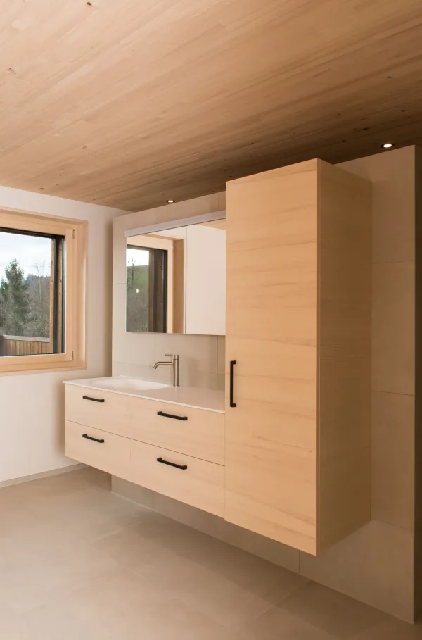 Innenansicht eines modernen Holzbaus mit sichtbaren Balken, natürlichen Holzoberflächen und massgefertigten Einbauten. Grosszügige Fensterflächen sorgen für viel Tageslicht und unterstreichen die warme, ruhige Wohnatmosphäre. Hochwertige Materialien und präzise Schreinerarbeiten prägen das Gesamtbild.