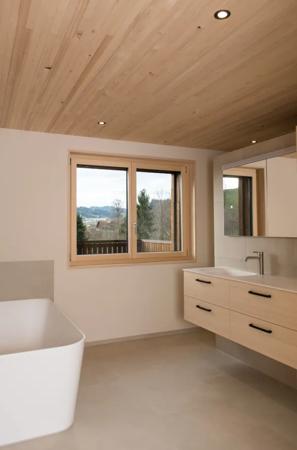 Innenansicht eines modernen Holzbaus mit sichtbaren Balken, natürlichen Holzoberflächen und massgefertigten Einbauten. Grosszügige Fensterflächen sorgen für viel Tageslicht und unterstreichen die warme, ruhige Wohnatmosphäre. Hochwertige Materialien und präzise Schreinerarbeiten prägen das Gesamtbild.