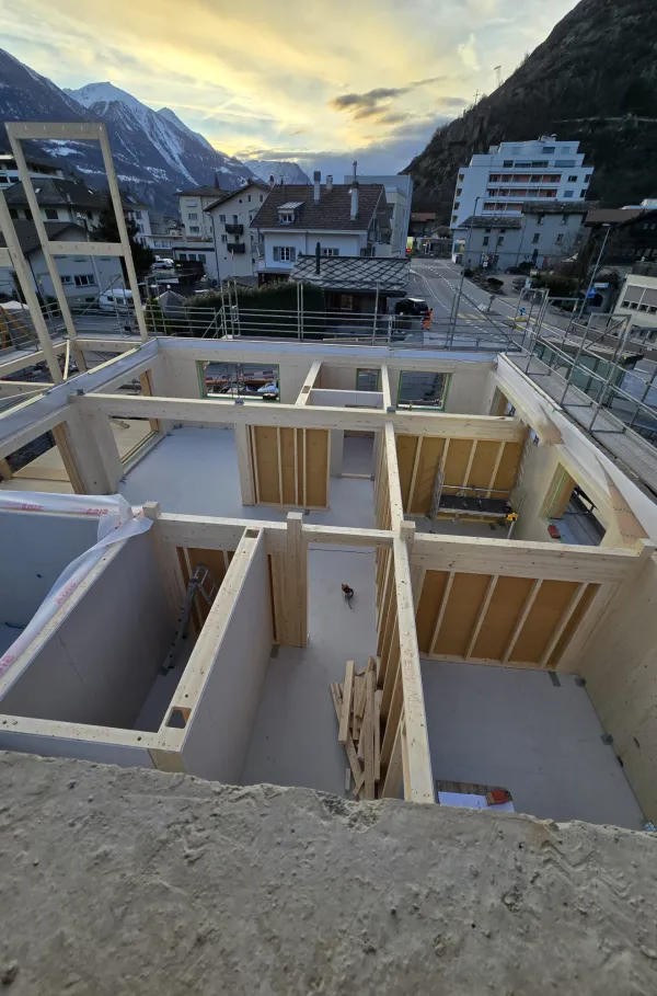 Zimmerleute arbeiten an der Holzrahmenkonstruktion eines mehrgeschossigen Gebäudes auf einer Baustelle. Die tragenden Wände aus Holz werden vor Ort montiert, während ein Baukran über dem Bauplatz steht. Im Hintergrund erstrecken sich Dorfhäuser und eine beeindruckende alpine Berglandschaft