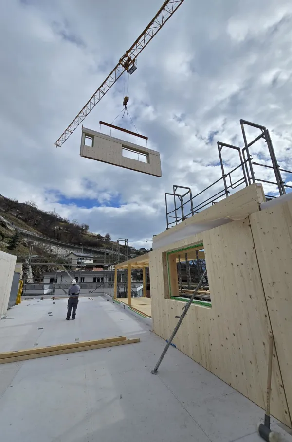 Zimmerleute arbeiten an der Holzrahmenkonstruktion eines mehrgeschossigen Gebäudes auf einer Baustelle. Die tragenden Wände aus Holz werden vor Ort montiert, während ein Baukran über dem Bauplatz steht. Im Hintergrund erstrecken sich Dorfhäuser und eine beeindruckende alpine Berglandschaft