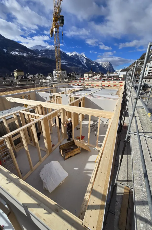 Zimmerleute arbeiten an der Holzrahmenkonstruktion eines mehrgeschossigen Gebäudes auf einer Baustelle. Die tragenden Wände aus Holz werden vor Ort montiert, während ein Baukran über dem Bauplatz steht. Im Hintergrund erstrecken sich Dorfhäuser und eine beeindruckende alpine Berglandschaft