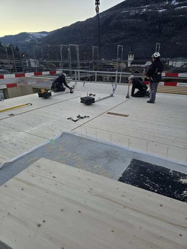 Zimmerleute arbeiten an der Holzrahmenkonstruktion eines mehrgeschossigen Gebäudes auf einer Baustelle. Die tragenden Wände aus Holz werden vor Ort montiert, während ein Baukran über dem Bauplatz steht. Im Hintergrund erstrecken sich Dorfhäuser und eine beeindruckende alpine Berglandschaft