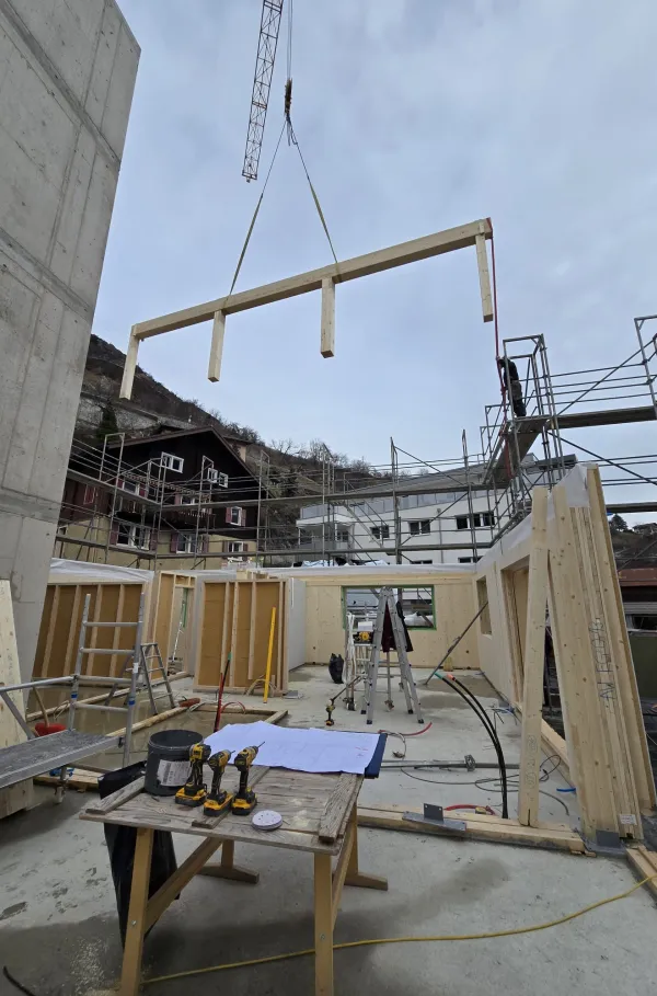 Zimmerleute arbeiten an der Holzrahmenkonstruktion eines mehrgeschossigen Gebäudes auf einer Baustelle. Die tragenden Wände aus Holz werden vor Ort montiert, während ein Baukran über dem Bauplatz steht. Im Hintergrund erstrecken sich Dorfhäuser und eine beeindruckende alpine Berglandschaft