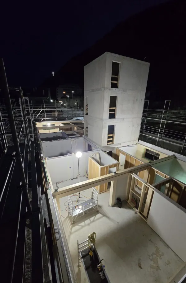 Zimmerleute arbeiten an der Holzrahmenkonstruktion eines mehrgeschossigen Gebäudes auf einer Baustelle. Die tragenden Wände aus Holz werden vor Ort montiert, während ein Baukran über dem Bauplatz steht. Im Hintergrund erstrecken sich Dorfhäuser und eine beeindruckende alpine Berglandschaft