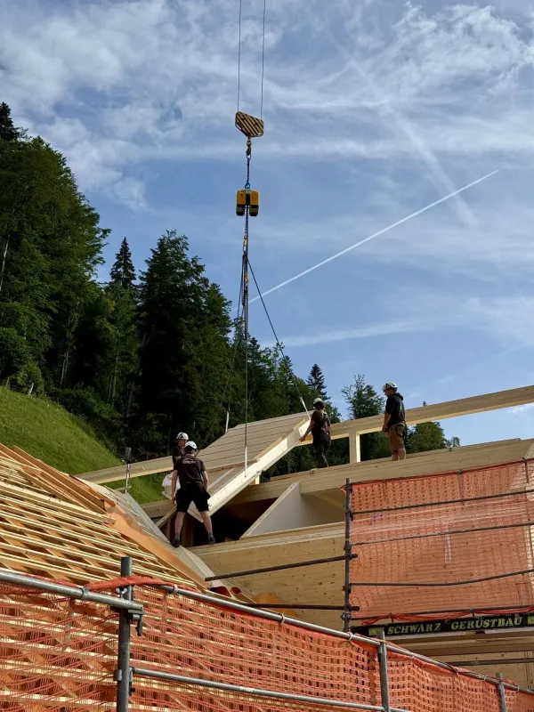 Zimmerleute montieren während der Aufrichtearbeiten eine vorgefertigte Holzkonstruktion auf einem Gebäude. Ein Kran hebt ein grosses Dachelement an seinen Platz, während mehrere Fachkräfte auf dem Dach die Montage begleiten.