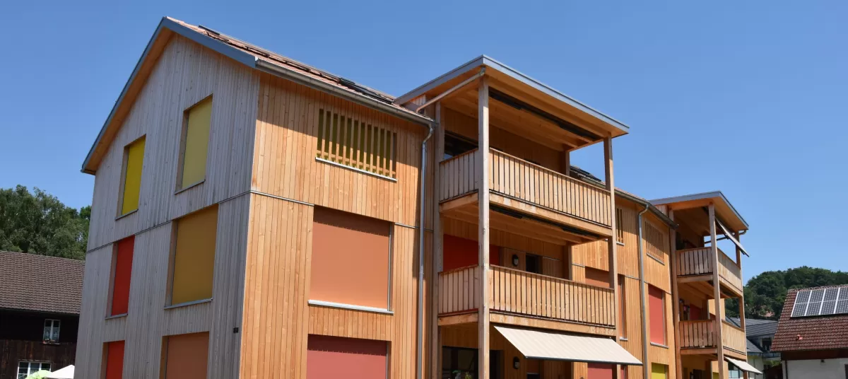 Mehrfamilienhaus aus Truberholz mit Balkon und Aussenplatz. Truberholzschallung stehend. Fabrkonzept bei den Storen. Naturbegrünung (Ökologischer Garten) um das Haus.