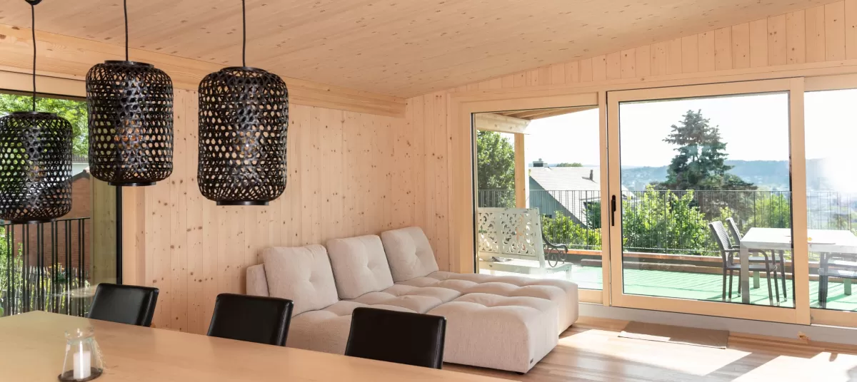 Wohnbereich mit Fensterfront, Truberholzwände ersichtlich, Decke Wände Boden aus Holz, wunderschön eingerichtet mit stylischen schwarzen Lampen, helles Sofa, Holztisch