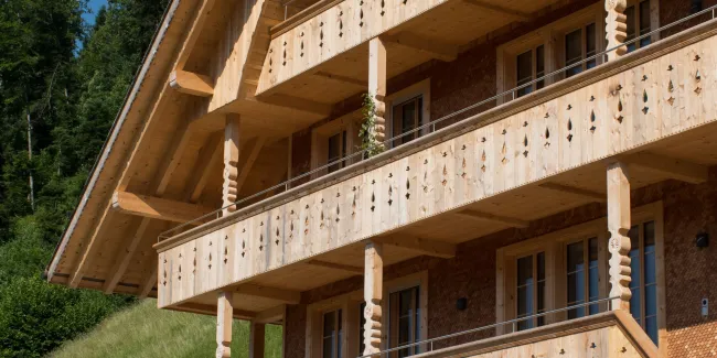 Mehrstöckiges Holzhaus im traditionellen Schweizer Stil mit kunstvoll verzierten Holzbalkonen und grosszügigem Dachüberstand. Die Fassade besteht aus hellem, massivem Holz, umgeben von einer grünen Hanglandschaft bei klarem Himmel.