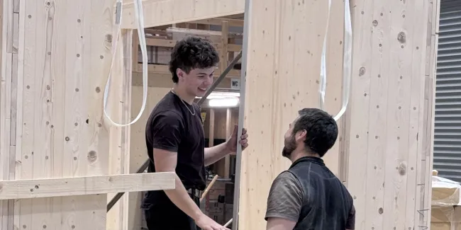 Zwei Mitarbeitende der Zimmerei Hirschi montieren in der Werkhalle ein vorgefertigtes Holzelement. Sie arbeiten an einer massiven Holzrahmenkonstruktion mit Fensteröffnung und stimmen sich dabei direkt vor Ort ab.