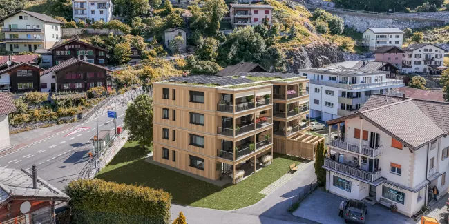 Visualisierung eines modernen Mehrfamilienhauses in Holzbauweise mit grossen Balkonen und begrüntem Dach mit Solaranlage. Das Gebäude fügt sich in eine alpine Dorflandschaft mit traditionellen Häusern, Strassen und einem bewaldeten Berghang im Hintergrund ein