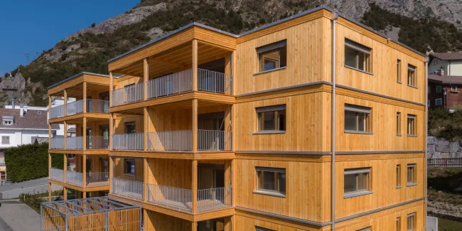 Aussenansicht eines modernen, viergeschossigen Mehrfamilienhauses in Holzbauweise vor einer felsigen Bergkulisse. Das Gebäude verfügt über eine vertikale Holzfassade sowie grosszügige, überdachte Balkone mit sichtbarer Holzkonstruktion und Metallgeländern.