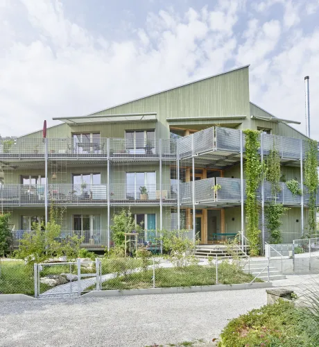 Aussenansicht eines modernen, mehrstöckigen Wohngebäudes in Holzbauweise mit hellgrüner Fassade. Das Haus verfügt über grosse Balkone mit Metallgeländern und Kletterpflanzen, die vertikal an den Fassadenelementen emporwachsen. Vor dem Gebäude liegt ein gepflegter Garten mit jungen Bäumen, Sträuchern und geschwungenen Wegen. Die Architektur wirkt offen und einladend, mit vielen Fenstern und einer klar strukturierten Formensprache. Im Vordergrund sind ein Kiesweg und ein Zaun sichtbar, während der Himmel leic