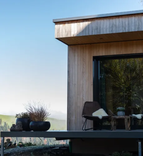 Aussenansicht eines modernen Holzhauses mit klarer Linienführung. Auf der erhöhten Terrasse stehen zwei Stühle mit Kissen und ein kleiner Tisch vor einer grossen Glasfront. Die Fassade besteht aus senkrechter Holzschalung, im Hintergrund sind Hügel, Bäume und eine ländliche Landschaft bei klarem Himmel zu sehen.