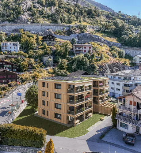 Visualisierung eines modernen Mehrfamilienhauses in Holzbauweise mit grossen Balkonen und begrüntem Dach mit Solaranlage. Das Gebäude fügt sich in eine alpine Dorflandschaft mit traditionellen Häusern, Strassen und einem bewaldeten Berghang im Hintergrund ein