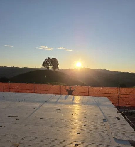 Blick über eine neu verlegte Holzdecke auf einer Baustelle bei Sonnenuntergang. Im Hintergrund steht ein Zimmermann am Gerüst vor der hügeligen Landschaft und hebt beide Arme in die Höhe, während die tief stehende Sonne über den Hügeln scheint.