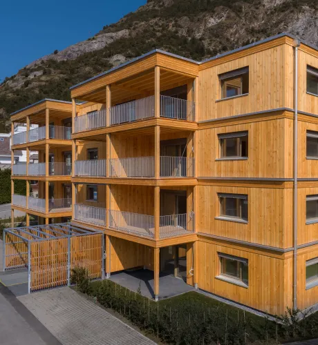 Aussenansicht eines modernen, viergeschossigen Mehrfamilienhauses in Holzbauweise vor einer felsigen Bergkulisse. Das Gebäude verfügt über eine vertikale Holzfassade sowie grosszügige, überdachte Balkone mit sichtbarer Holzkonstruktion und Metallgeländern.