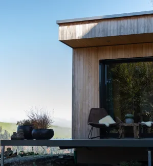 Aussenansicht eines modernen Holzhauses mit klarer Linienführung. Auf der erhöhten Terrasse stehen zwei Stühle mit Kissen und ein kleiner Tisch vor einer grossen Glasfront. Die Fassade besteht aus senkrechter Holzschalung, im Hintergrund sind Hügel, Bäume und eine ländliche Landschaft bei klarem Himmel zu sehen.