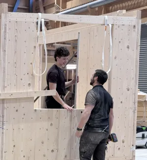 Zwei Mitarbeitende der Zimmerei Hirschi montieren in der Werkhalle ein vorgefertigtes Holzelement. Sie arbeiten an einer massiven Holzrahmenkonstruktion mit Fensteröffnung und stimmen sich dabei direkt vor Ort ab.