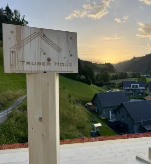 Zimmerleute montieren während der Aufrichtearbeiten eine vorgefertigte Holzkonstruktion auf einem Gebäude. Ein Kran hebt ein grosses Dachelement an seinen Platz, während mehrere Fachkräfte auf dem Dach die Montage begleiten.