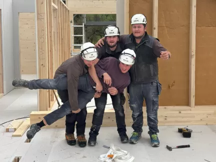 Vier Zimmerleute posieren lachend auf der Baustelle eines Holzhauses. Die fröhliche Stimmung zeigt den Teamgeist und die Freude am Handwerk bei der Zimmerei Hirschi AG.