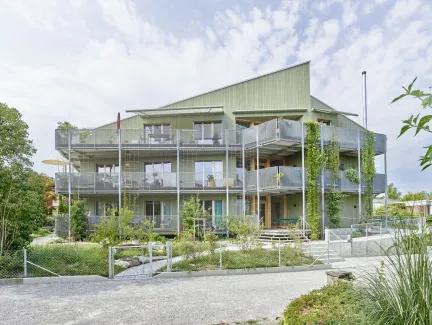 Aussenansicht eines modernen, mehrstöckigen Wohngebäudes in Holzbauweise mit hellgrüner Fassade. Das Haus verfügt über grosse Balkone mit Metallgeländern und Kletterpflanzen, die vertikal an den Fassadenelementen emporwachsen. Vor dem Gebäude liegt ein gepflegter Garten mit jungen Bäumen, Sträuchern und geschwungenen Wegen. Die Architektur wirkt offen und einladend, mit vielen Fenstern und einer klar strukturierten Formensprache. Im Vordergrund sind ein Kiesweg und ein Zaun sichtbar, während der Himmel leic
