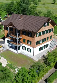 Luftaufnahme eines frisch renovierten, mehrstöckigen Holzhauses im traditionellen Stil mit brauner Schindelfassade und grünen Fensterläden. Das Gebäude steht inmitten einer ländlichen Umgebung mit Wiesen, Bäumen und benachbarten Wohn- und Wirtschaftsbauten. Vor dem Haus befindet sich ein neu angelegter Vorplatz, während rundherum noch Bereiche der Gartengestaltung und Bauarbeiten sichtbar sind. Im Hintergrund erstreckt sich eine grüne Hügellandschaft mit verstreuten Obstbäumen und einigen modernen Wohnhäuse