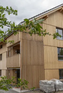 Aussenansicht eines neu errichteten Mehrfamilienhauses in moderner Holzbauweise mit senkrechter Holzfassade und Satteldach. Balkone und Fassadenelemente sind aus Holz gestaltet. Im Vordergrund sind Pflanzen und Baustellenmaterialien zu sehen, im Hintergrund ein Wohnquartier unter bewölktem Himmel.