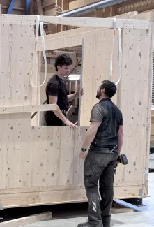 Zwei Mitarbeitende der Zimmerei Hirschi montieren in der Werkhalle ein vorgefertigtes Holzelement. Sie arbeiten an einer massiven Holzrahmenkonstruktion mit Fensteröffnung und stimmen sich dabei direkt vor Ort ab.