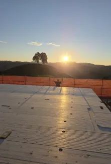 Blick über eine neu verlegte Holzdecke auf einer Baustelle bei Sonnenuntergang. Im Hintergrund steht ein Zimmermann am Gerüst vor der hügeligen Landschaft und hebt beide Arme in die Höhe, während die tief stehende Sonne über den Hügeln scheint.