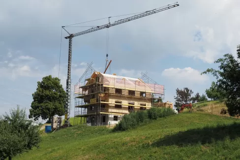 Ein mehrstöckiges Holzhaus im Bau, umgeben von Gerüsten und einem Baukran, steht auf einer grünen Anhöhe unter blauem Himmel mit einigen Wolken.