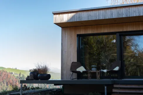 Aussenansicht eines modernen Holzhauses mit klarer Linienführung. Auf der erhöhten Terrasse stehen zwei Stühle mit Kissen und ein kleiner Tisch vor einer grossen Glasfront. Die Fassade besteht aus senkrechter Holzschalung, im Hintergrund sind Hügel, Bäume und eine ländliche Landschaft bei klarem Himmel zu sehen.