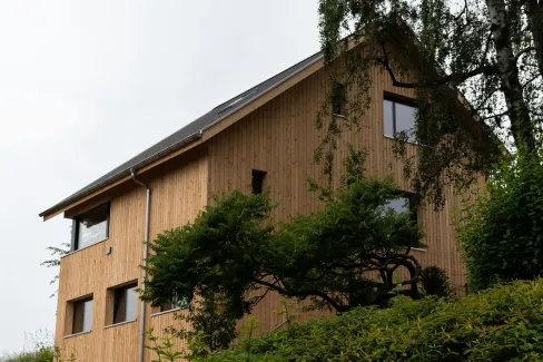 Aussenansicht eines modernen Einfamilienhauses in Holzbauweise mit Satteldach und dunkler Dacheindeckung. Das Gebäude verfügt über eine Terrasse und einen Balkon mit Metallgeländer. Es steht an leichter Hanglage, umgeben von Grünflächen und angrenzenden Wohnhäusern, vor bewaldetem Hintergrund.